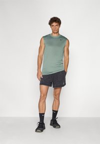 Grøn ærmeløs træningsskjorte, sorte shorts med logo, sorte træningssko og sorte sokker. Glat stof, tætsiddende design og afslappet stil.