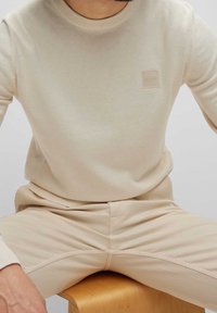 Beige långärmad tröja med ribbad halsringning och en liten, fyrkantig, präglad "BOSS"-logotyp på bröstet. Mjuk textur, figurnära design.