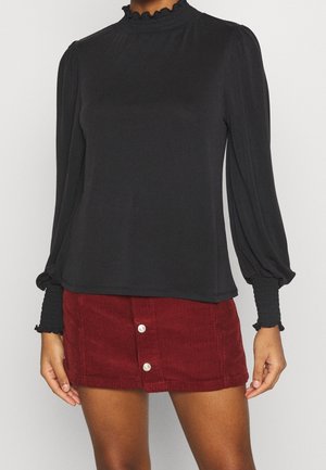 Woman wears a black long-sleeve top with smocked cuffs and collar, paired with a red corduroy mini skirt with front buttons.