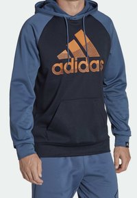 Navy and blue hooded sweatshirt with kangaroo pocket, featuring an orange Adidas logo. Soft fabric with a relaxed fit and contrasting raglan sleeves.