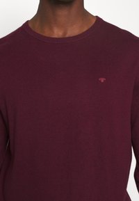 Burgundy knitted sweater with a round neckline. Features a small, tonal logo on the chest. Texture appears smooth and fitted.