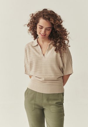 Kobieta o kręconych włosach, ubrana w beżowy, krótki sweter z krótkim rękawem i oliwkowo-zielone spodnie, uśmiechająca się z zamkniętymi oczami, trzymająca ręce za plecami.