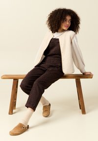 Krämfärgad shearlingväst över en ljusgrå långärmad tröja, parat med mörkbruna leopardmönstrade hängselbyxor och beige klackar med rem.