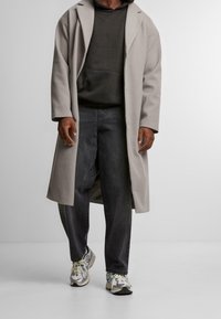 Světle šedý dlouhý kabát s klopou, kombinovaný s černou mikinou s kapucí, tmavými džínami a stříbrnými teniskami s barevnými akcenty.