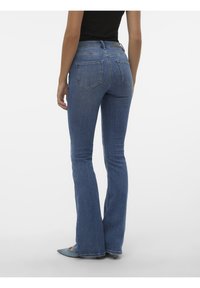 Femme portant un jean bootcut en denim bleu et un haut noir, debout de dos face à la caméra sur un fond blanc uni.