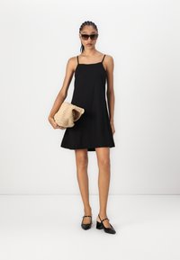Rochie neagră fără mâneci, cu decolteu drept, siluetă în formă de A; modelul ține o poșetă din paie împletită; purtată cu pantofi cu toc negri.
