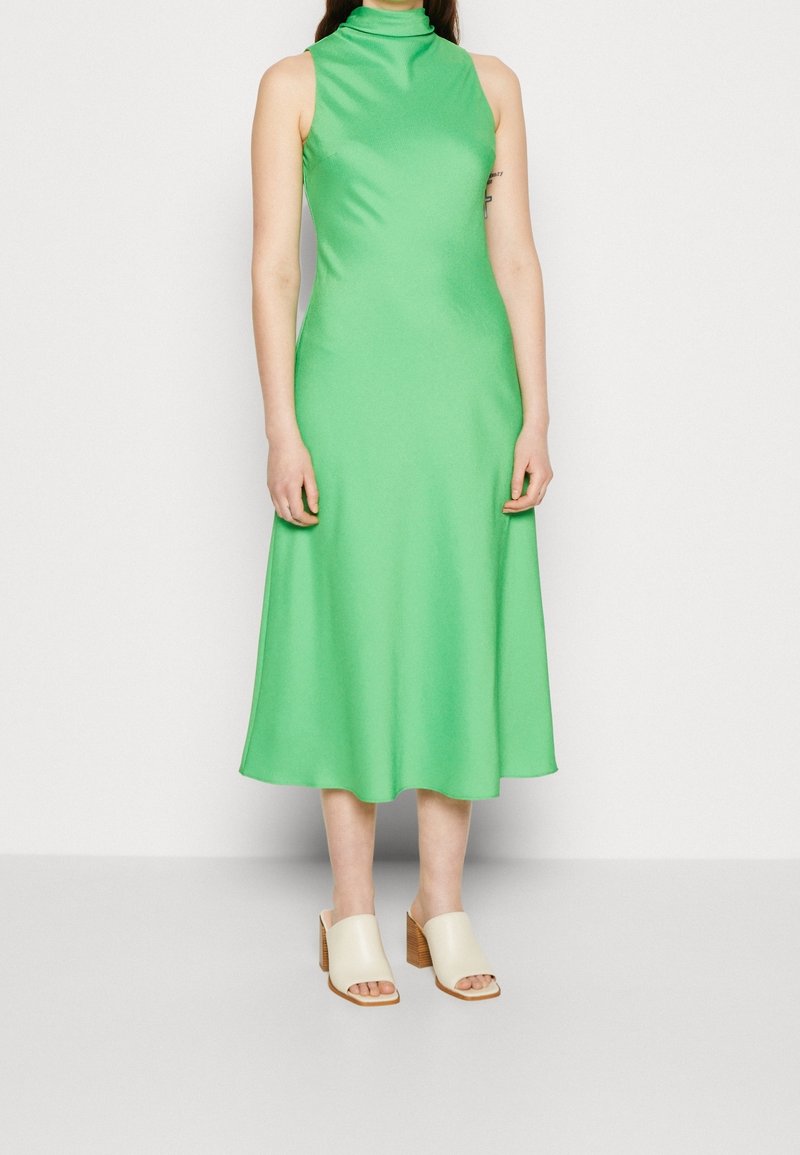 Zielona sukienka midi bez rękawów z wysokim kołnierzem, gładką teksturą i rozkloszowanym dołem. Do tego beżowe klapki na obcasie z drewnianym słupkiem.