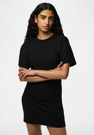 Femme aux longs cheveux bouclés debout sur un fond uni, portant une robe noire courte à manches courtes, bras croisés, regardant devant elle.