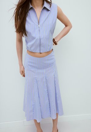 Crop top sans manches à rayures verticales bleu et blanc avec fermeture éclair, associé à une jupe midi plissée taille haute assortie et des bracelets dorés.