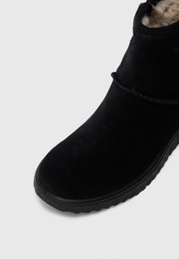 Botín negro de ante hasta el tobillo con una suela de goma texturizada, puntera suavemente redondeada y un acento cosido alrededor de la parte superior. Forrado con un material similar a la piel de lana.
