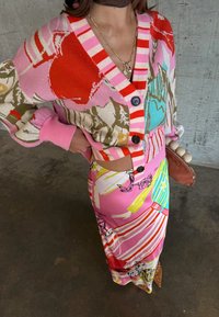 Γυναίκα που φοράει πολύχρωμο, μοτίβο ζακέτα με κουμπιά και μακριά φούστα που ταιριάζει, κρατώντας καφέ φάκελο με στρογγυλές διακοσμήσεις.