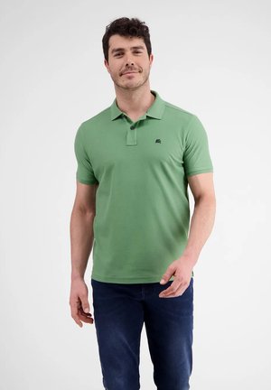Man standing, wearing light green short-sleeve polo shirt and dark blue jeans, against plain white background.
