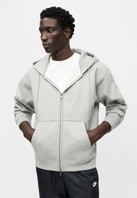 Γκρι ανοιχτόχρωμο φούτερ με φερμουάρ, από μαλακό ύφασμα, με μπροστινή τσέπη, ρυθμιζόμενο σκουφάκι με κορδόνι και ριμπ μανσέτες. Φορώντας το πάνω από ένα λευκό t-shirt.