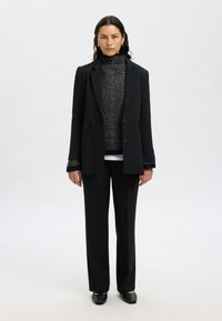 Črn krojen blazer z zarezanim ovratnikom, kombiniran s črno-belim vzorčastim puloverjem z visokim ovratnikom, črnimi hlačami in črnimi natikači.