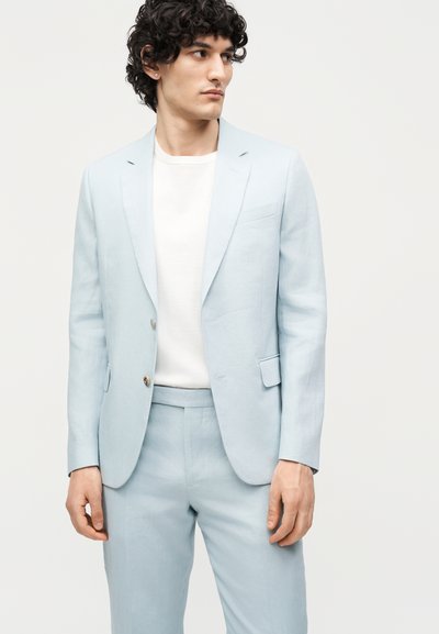 Νέος άνδρας με σγουρά μαλλιά, που φορά ανοιχτό μπλε κοστούμι και λευκό πουκάμισο, κοιτάζει προς τα δεξιά σε απλό φόντο.