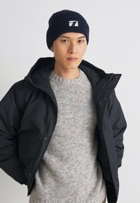 Veste zippée noire portée sur un pull texturé gris clair et un bonnet en tricot bleu marine avec un logo carré blanc. Le modèle pose devant un fond uni.