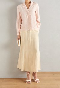 Lichtroze linnen blouse met lange mouwen en V-hals, gecombineerd met een beige, gestructureerde maxi-rok en witte sandalen met bandjes, met een kleine clutch in de hand.