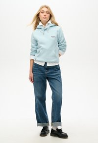 Sudadera azul clara con cordones y bolsillo frontal, superpuesta a una camisa blanca con cuello. Combinada con vaqueros azul claros de corte holgado y zapatos negros.