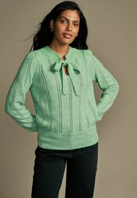 Pull en tricot vert avec un col à nouer, présentant un côtes verticales et des détails ajourés. Assorti avec un pantalon noir, mettant en valeur une silhouette ajustée.