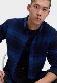 Blå ternet skjorte med knapper i uldblanding, med brystlomme, klassisk krave og sorte accenter. Modellen er stylet med en sort skjorte under.