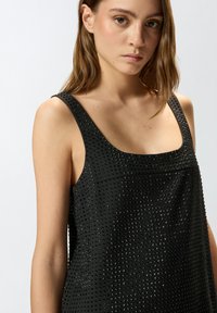 Top negro sin mangas con una superficie texturizada decorada con pequeños clavos, tirantes anchos y escote cuadrado, que ofrece una silueta entallada pero relajada.