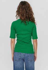 Person med lockigt brunt hår bär en grön t-shirt med halv ärm och blå denimjeans, vänd bort mot en enkel ljus bakgrund.