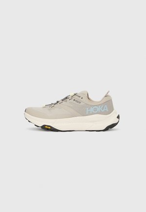 Beige athletic shoe with Gore-Tex upper, round toe, black Vibram sole, and reflective laces. Features light blue logo on the side.