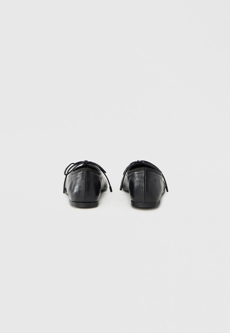 Chaussures plates en cuir noir avec un bout arrondi, dotées d'un petit talon et d'un nœud décoratif à l'arrière. Texture lisse avec un design minimaliste.