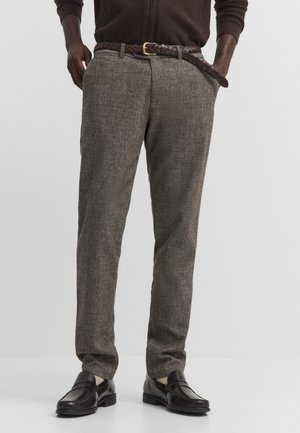Mann trägt strukturierte graue Hose mit geflochtenem braunem Gürtel, dunkelbraune Schuhe und ein dunkelbraunes Langarmoberteil, Hände in den Taschen.
