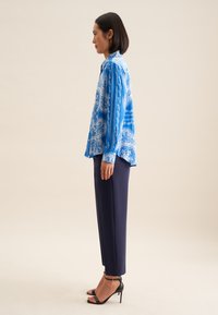 Blusa blu con motivo, in tessuto morbido, abbottonata, con maniche lunghe. Indossata con pantaloni blu navy e sandali con tacco neri, in vista laterale.