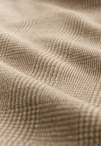 Tejido beige con un patrón texturizado que presenta líneas y puntos visibles, creando una sutil apariencia de cuadrícula en toda su superficie.