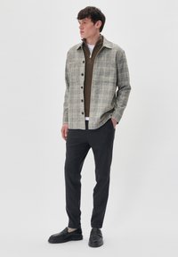 Cămașă cu carouri în gri deschis și bej, suprapusă peste un pulover cu fermoar maro, asortată cu pantaloni în dungi închise și pantofi negri.