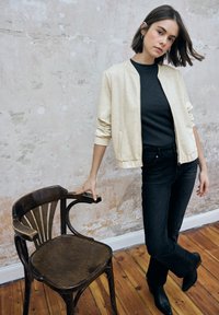 Chaqueta bomber crema con textura moteada, top negro acanalado, jeans negros ajustados y botines negros, posando junto a una silla de madera.