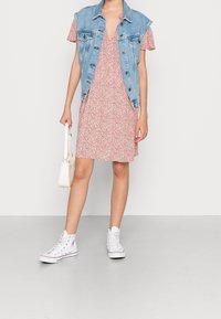 Veste en denim sur une robe courte à motifs floraux roses et blancs, associée à des baskets montantes blanches. Petit sac à main blanc simple.