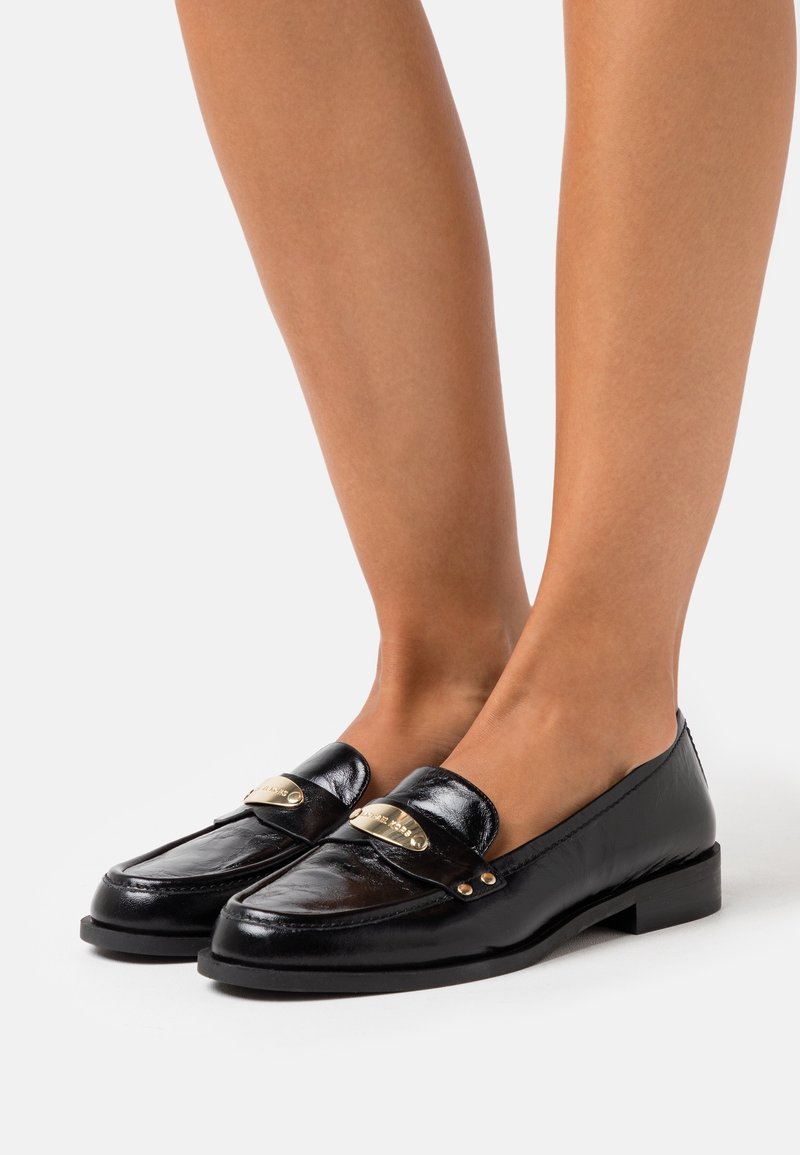 Mocassins en cuir noir avec un bout arrondi, présentant un accent doré et un petit talon carré. Texture lisse avec des détails de couture.