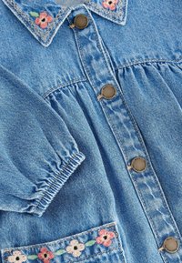 Vêtement en denim bleu avec boutons en métal, manches à poignets élastiques et fleurs roses et blanches brodées sur le col et les bords des poches.