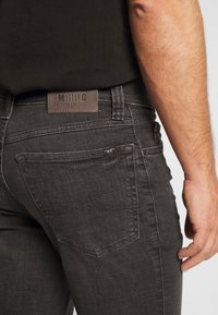 Jeans de mezclilla gris oscuro con un parche de cuero marrón etiquetado "MUSTANG True Denim", bolsillos traseros con detalles de costura. Textura suave visible.