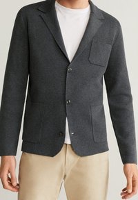 Uomo che indossa un blazer grigio scuro con bottoni e tre tasche sopra una t-shirt bianca e pantaloni beige su uno sfondo chiaro.