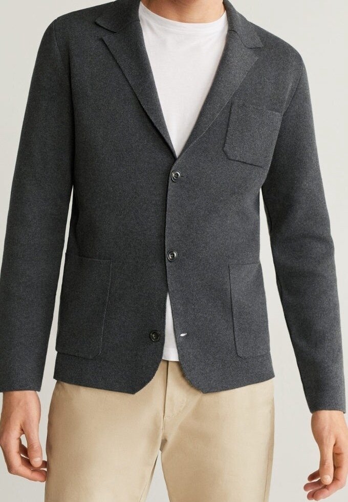 Uomo che indossa un blazer grigio scuro con bottoni e tre tasche sopra una t-shirt bianca e pantaloni beige su uno sfondo chiaro.
