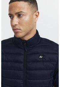 Mørkeblå polstret jakke med høy krage og glidelås foran. Har horisontal søm og en liten logo på venstre side. Glatt tekstur.