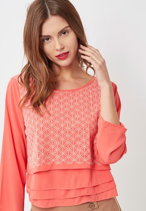 Blusa coral de manga larga con bordado geométrico blanco en la parte superior y tejido en capas en el dobladillo. Textura suave.