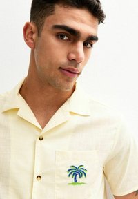 Camicia a maniche corte gialla chiaro con colletto tradizionale, caratterizzata da una tasca frontale e ricami di palme blu e verdi. Dettagli con bottoni dorati.