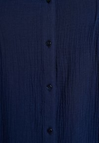 Predná časť košele z tmavomodrej textúrovanej tkaniny so západkami s piatimi čiernymi gombíkmi rovnomerne rozmiestnenými vertikálne.