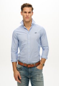 Camicia a maniche lunghe a righe blu e bianche con colletto a punte. Presenta un piccolo logo sul petto, abbinata a jeans di denim e una cintura marrone.