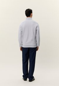 Uomo in piedi di spalle che indossa un pullover grigio chiaro a maniche lunghe e pantaloni blu navy a gamba dritta con scarpe nere.