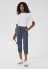 Granatowe spodenki  cropped z elastycznym paskiem i sznurkiem, w połączeniu z dopasowaną białą koszulką. Model trzyma w dłoni okulary przeciwsłoneczne i ma na sobie białe trampki.