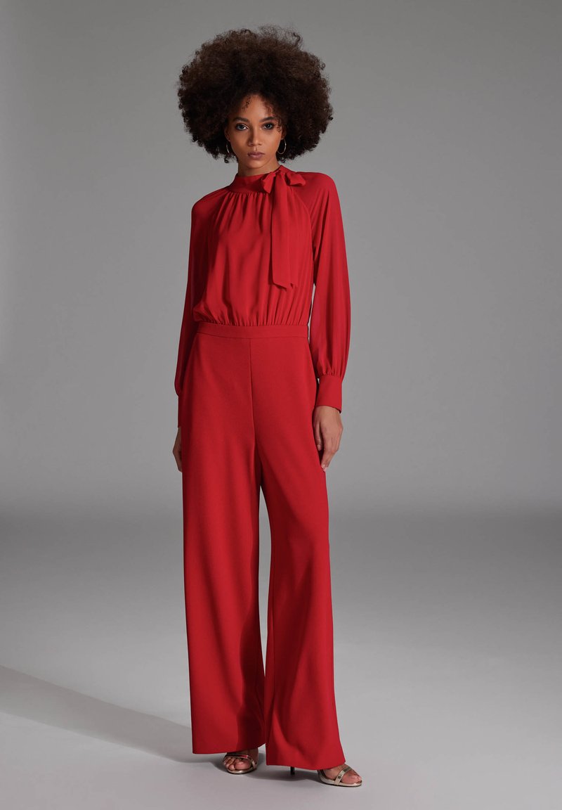 Vrouw met afro-haarstijl, gekleed in een rode blouse met lange mouwen en strik, in wijde broek, staand tegen een grijze achtergrond.