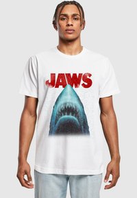 T-shirt en coton blanc avec un graphique d'un requin sous le mot "JAWS" en rouge, sur un arrière-plan texturé bleu et avec des détails visibles.