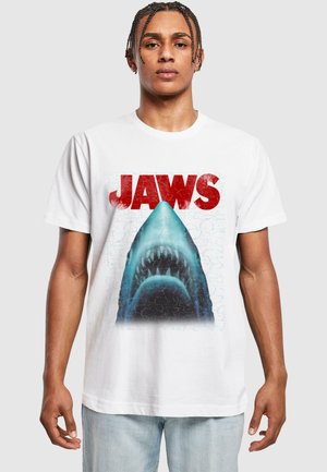 Hvid bomulds T-shirt med en grafik af en haj under ordet "JAWS" i rød, med en tekstureret blå baggrund og synlige detaljer.
