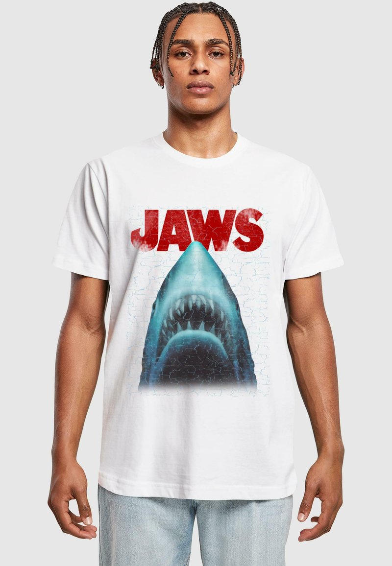 T-shirt en coton blanc avec un graphique d'un requin sous le mot "JAWS" en rouge, sur un arrière-plan texturé bleu et avec des détails visibles.